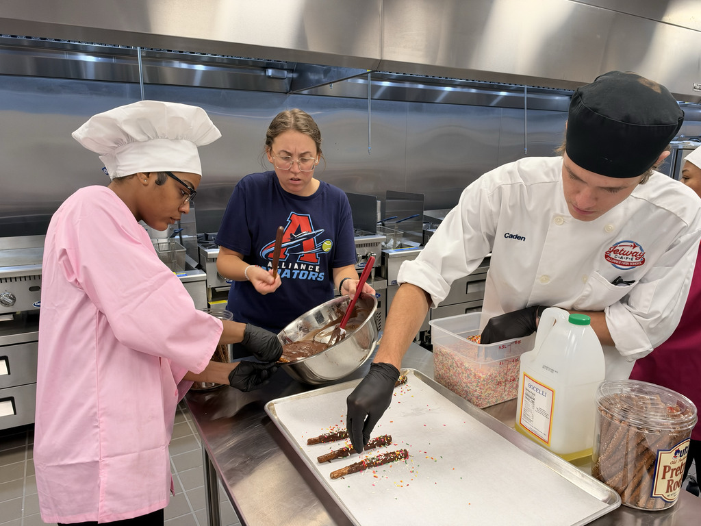 Two students dressed in chef coats and hats put sprinkles on pretzel rods they covered in chocolate