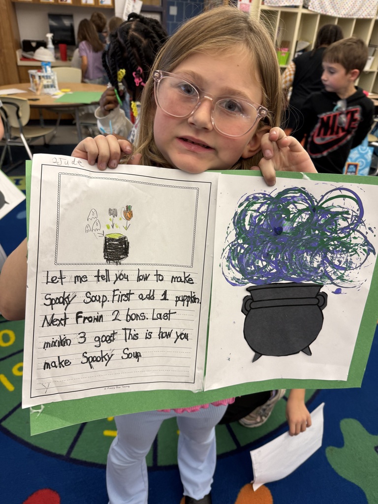 A student holds up the essay she wrote and the picture of her spooky soup she created