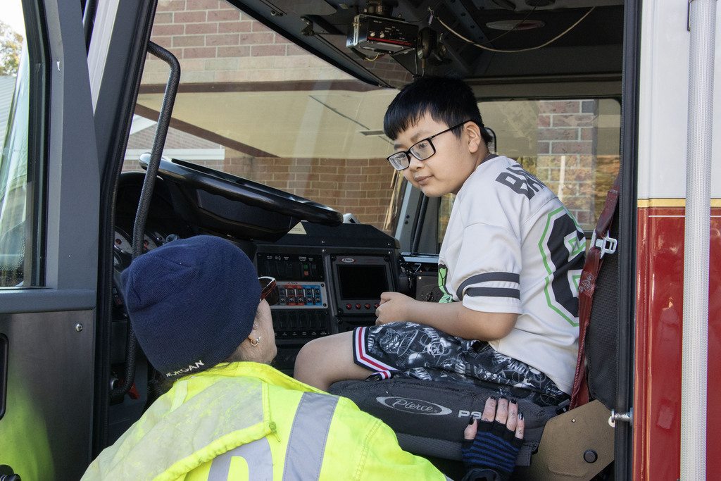 A student sits in the drivers seat of the fire truck while the firefighter stands beside him