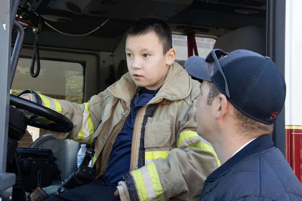 A student wearing a fire fighter coat sits in the drivers seat of the fire truck while the firefighter stands beside him