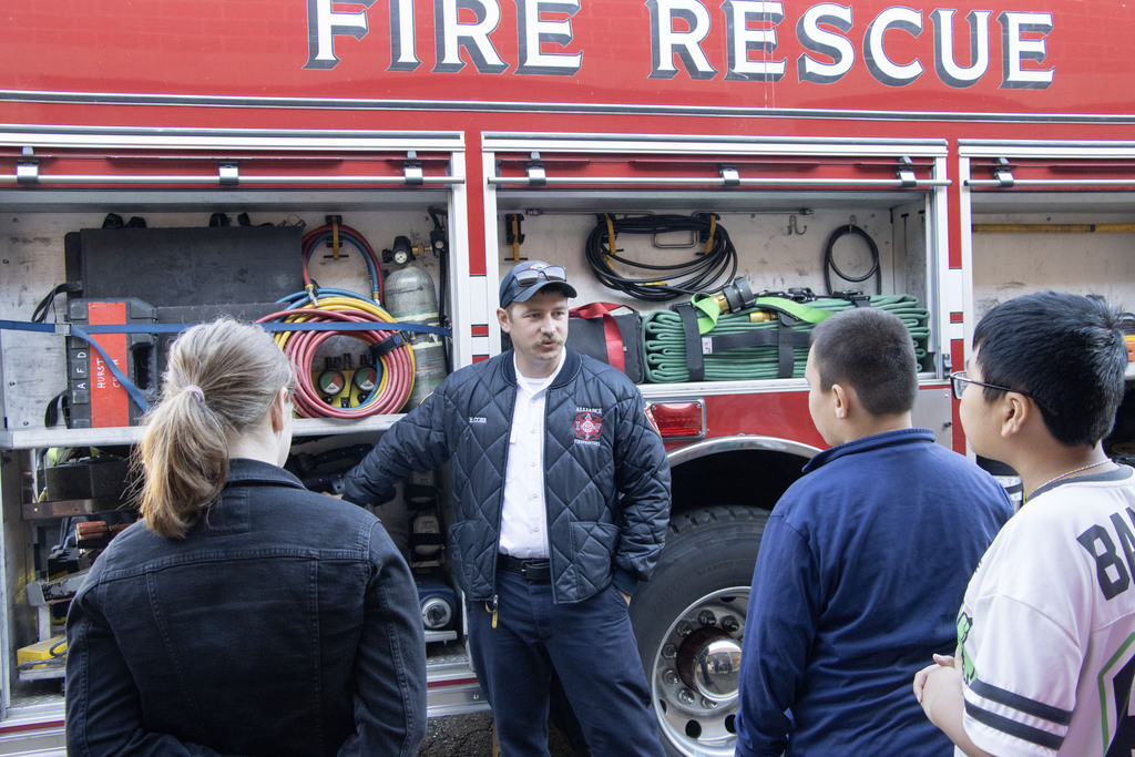 A fire fighter points to something on the  firetruck while students watch