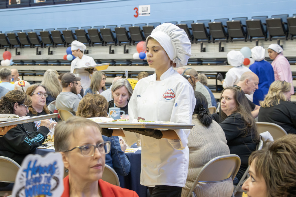 A student wearing a chefs coat and hat and holding a tray walks between crowded tables at the banquet
