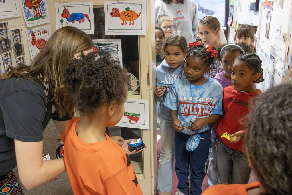 A student in an orange shirt presses a button that says "trick or treat" while students in the class room wait to pass out candy