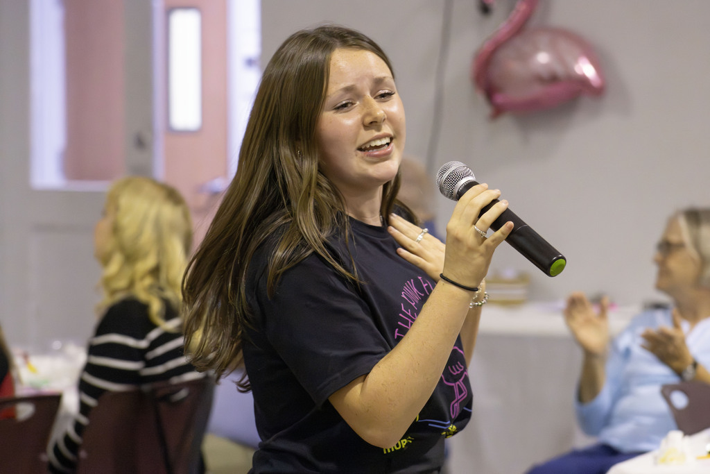 A student from the cast holds a microphone and sings while standing among the audience