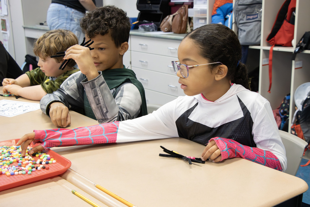a girl dressed as spider woman uses beads and pipe cleaners to create a spider