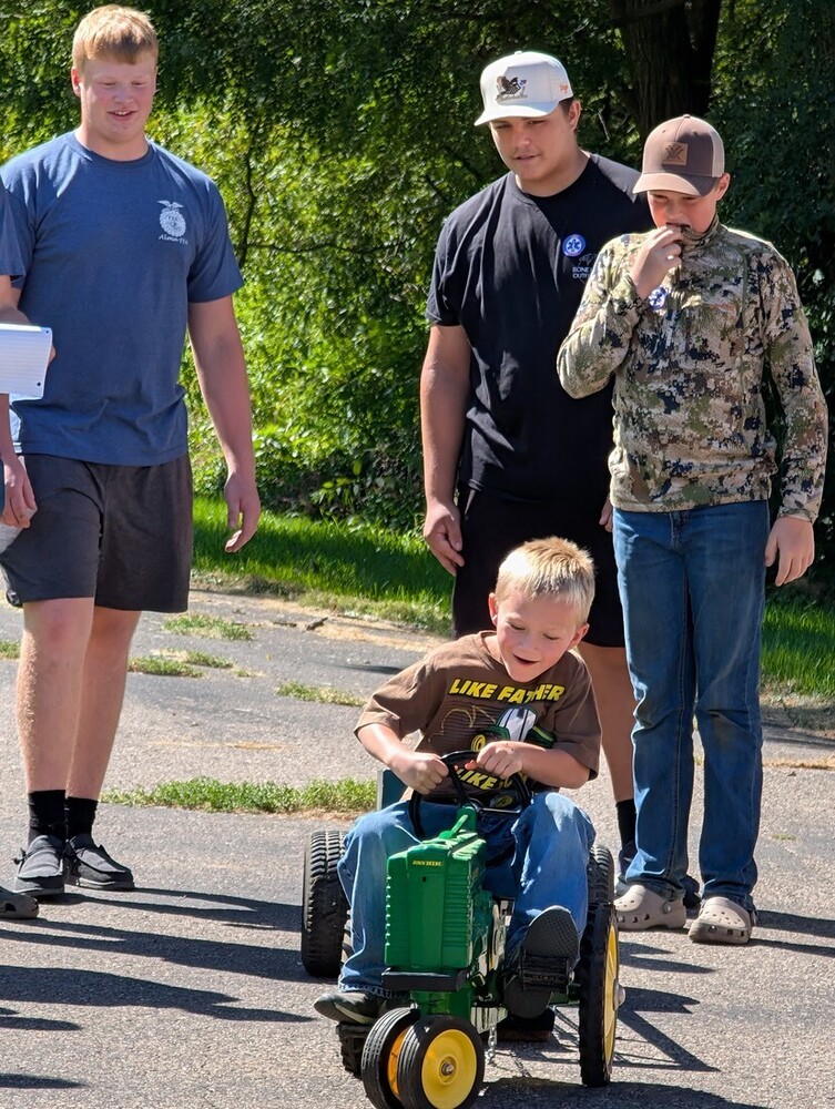 A young child sits on a small green John Deere tractor during a community event, surrounded by several older children and teens. One person wears a camouflage hunting outfit, while another wears a black shirt with an EMS-related logo. They stand outdoors on a sunny day with trees and a residential street in the background.