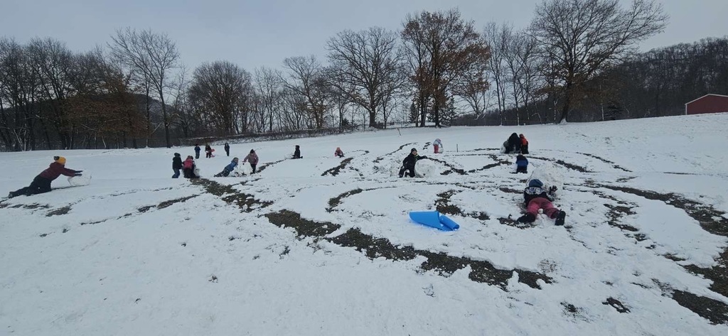Several children in winter clothing collaborate to roll snowballs across a snow-covered hill as they build a snowman. The hillside is lightly covered with snow, with some exposed grass visible. Bare trees and brush form a wooded background beneath a cloudy winter sky.