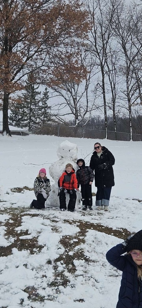 Several children and a teacher in winter clothing posing with the finished snowman. One student off to the side in black jacket in bottom of photo, one with a pink hat and white and black jacket, one with a bright orange and black jacket, one with a black jack and a teacher in a black jacket pose in front of the snowman. The hillside is lightly covered with snow, with some exposed grass visible. Bare trees and brush form a wooded background beneath a cloudy winter sky.
