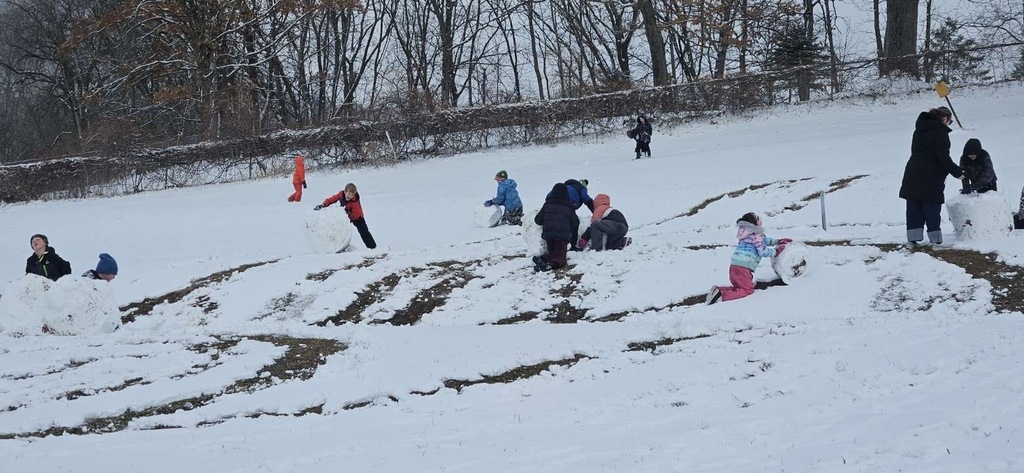 Several children in winter clothing collaborate to assemble a snowman across a snow-covered hill. In the foreground, one child in a blue and black jacket and another in a pink and gray jacket push the snowball together. Other children, including one in a bright orange snowsuit and another crouching farther up the hill, play in the snow nearby. The hillside is lightly covered with snow, with some exposed grass visible. Bare trees and brush form a wooded background beneath a cloudy winter sky.