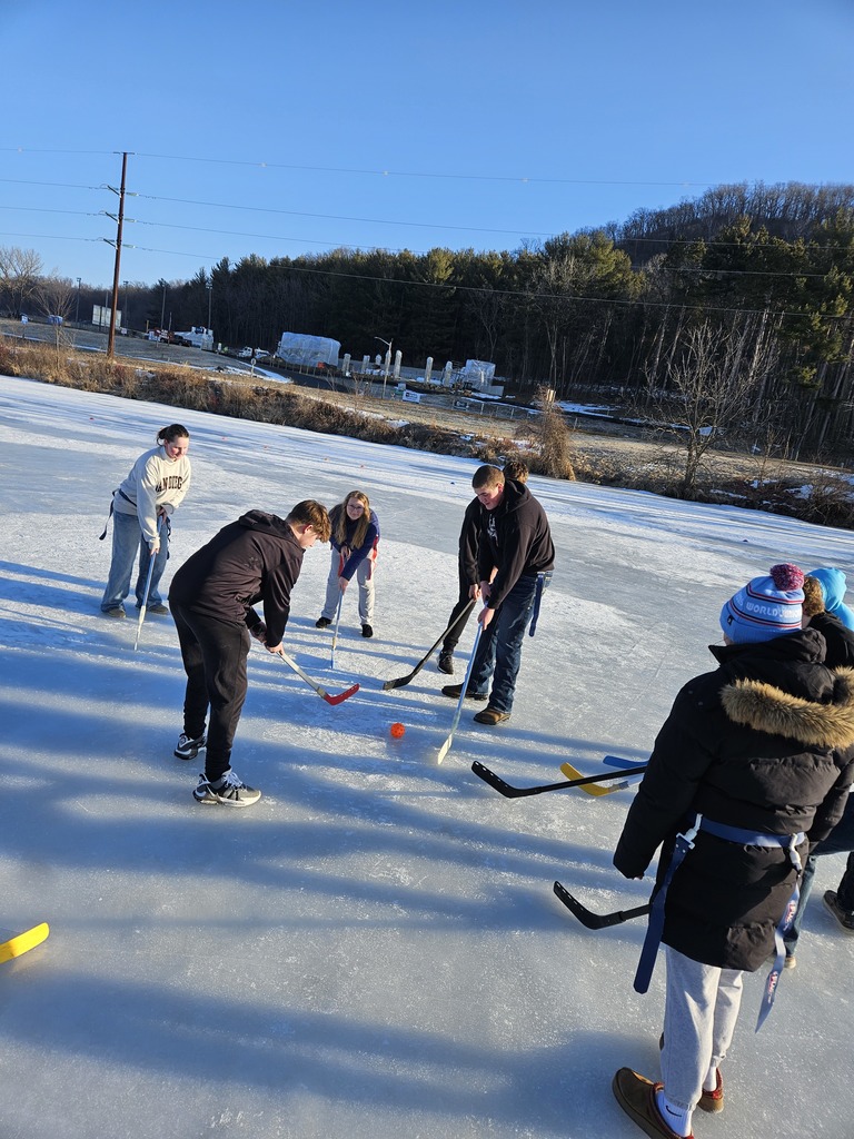 The image contains students outside on the ice. They are playing hockey with a wiffle ball and plastic hockey sticks.