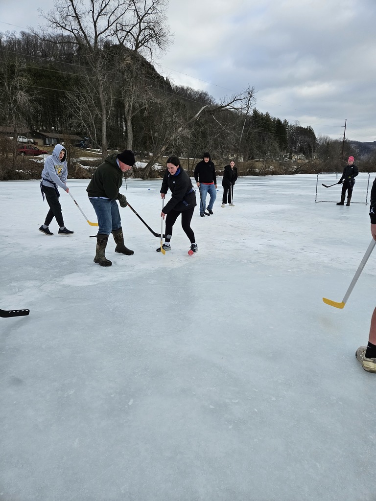 The image contains students outside on the ice. They are playing hockey with a wiffle ball and plastic hockey sticks.