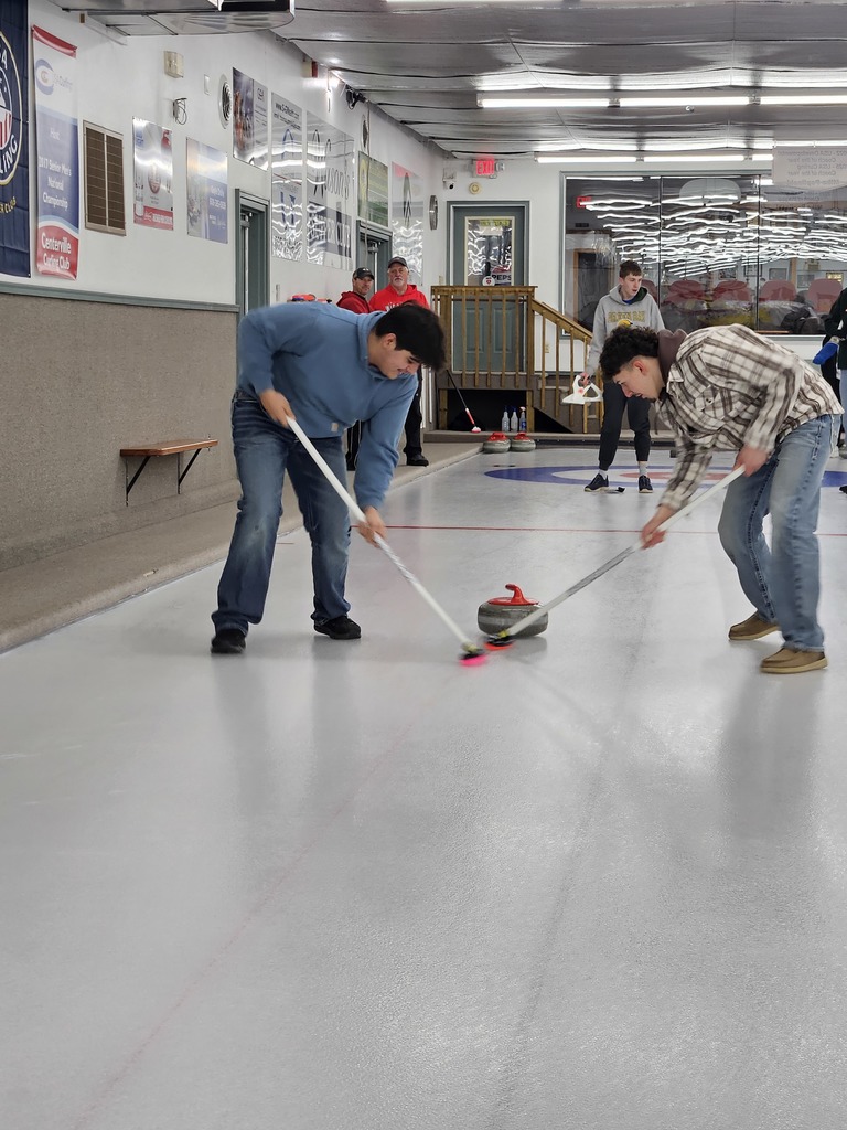 Two people sweep the ice with curling brooms as a red curling stone slides down an indoor rink, while others watch from behind.