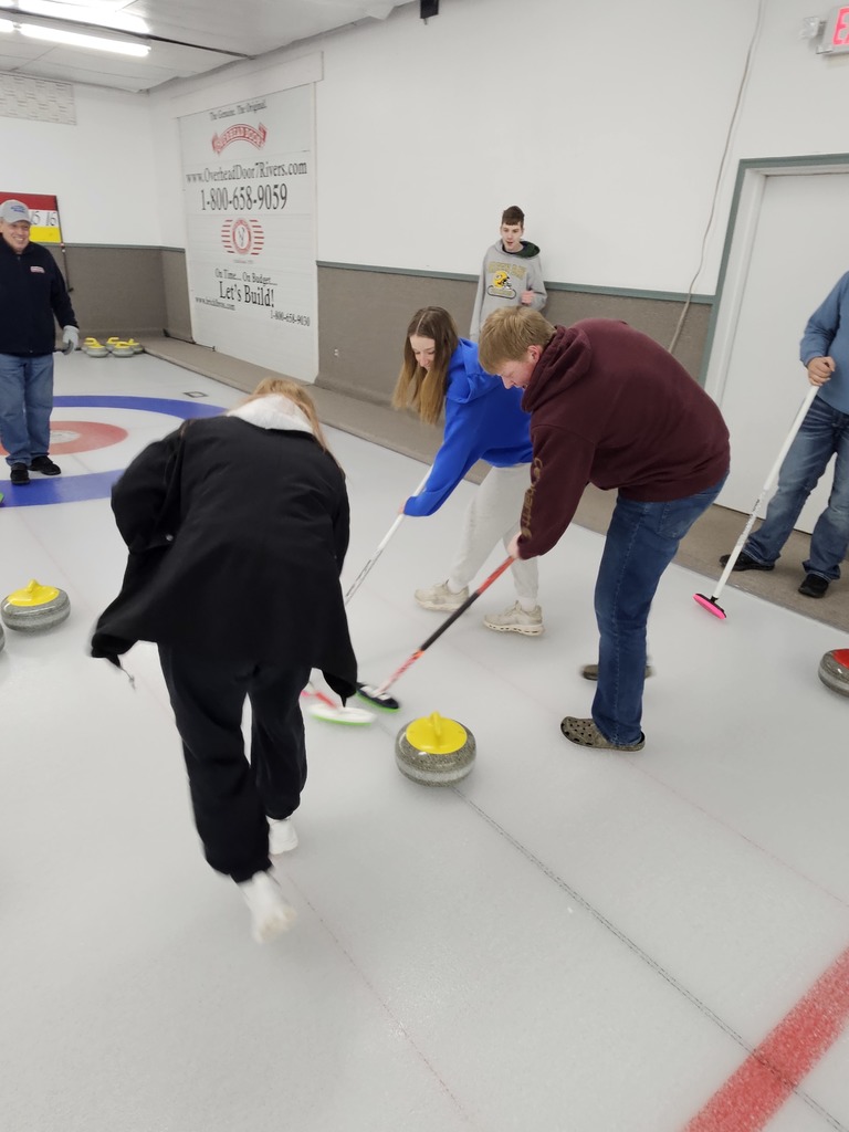 Several people play curling indoors, sweeping the ice with brooms as a yellow curling stone slides toward the target area while others watch nearby.