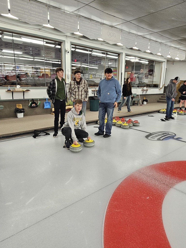 A group of young people stand on an indoor curling rink as one person crouches to release two yellow curling stones while others watch and wait nearby.