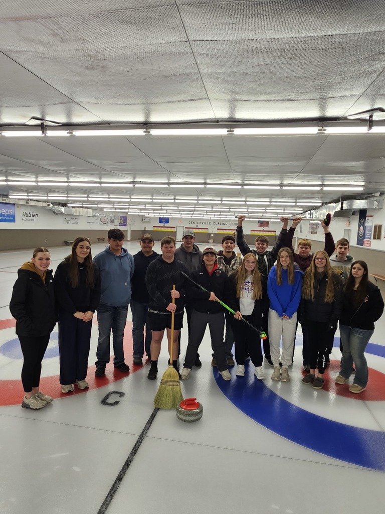 A group of people pose together on an indoor curling rink, standing on the target rings with curling brooms and a red curling stone in front of them.