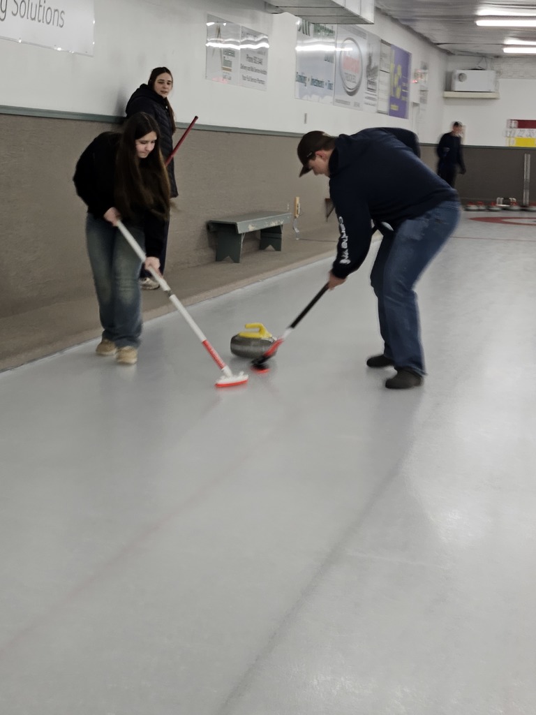 Two people sweep the ice with curling brooms as a yellow curling stone moves down an indoor curling rink, while another person watches from the side.