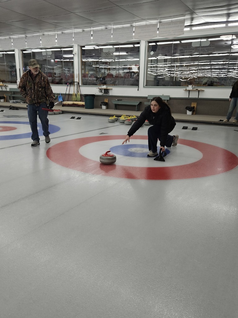 A person slides a red curling stone toward the target rings on an indoor curling rink while another person stands nearby watching.
