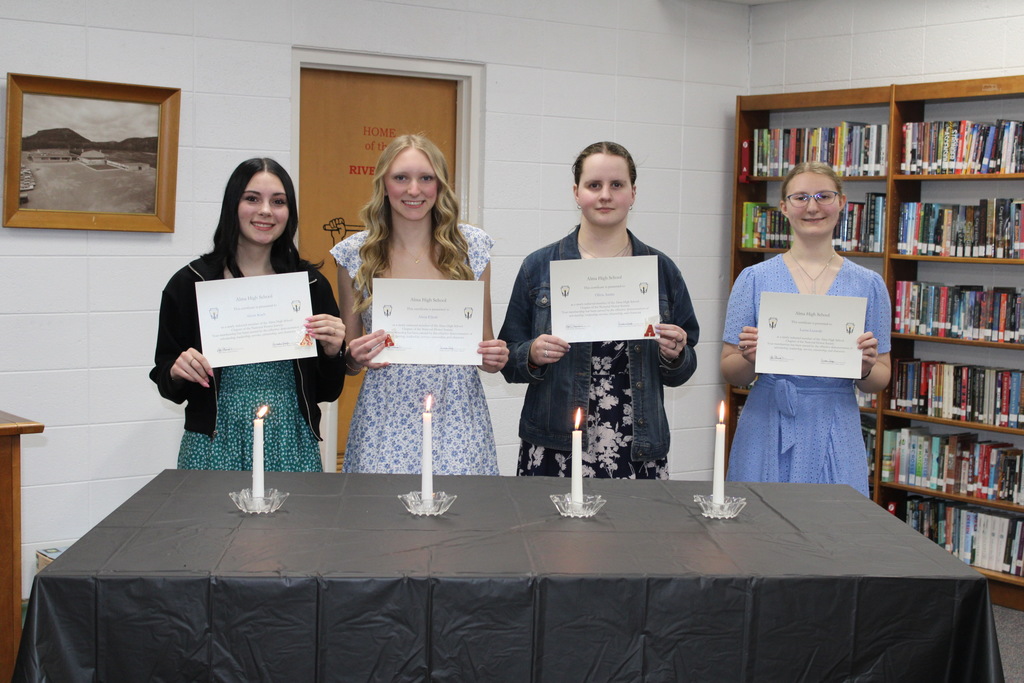 Four students stand behind a table with lit candles in a library, holding certificates during a recognition ceremony.