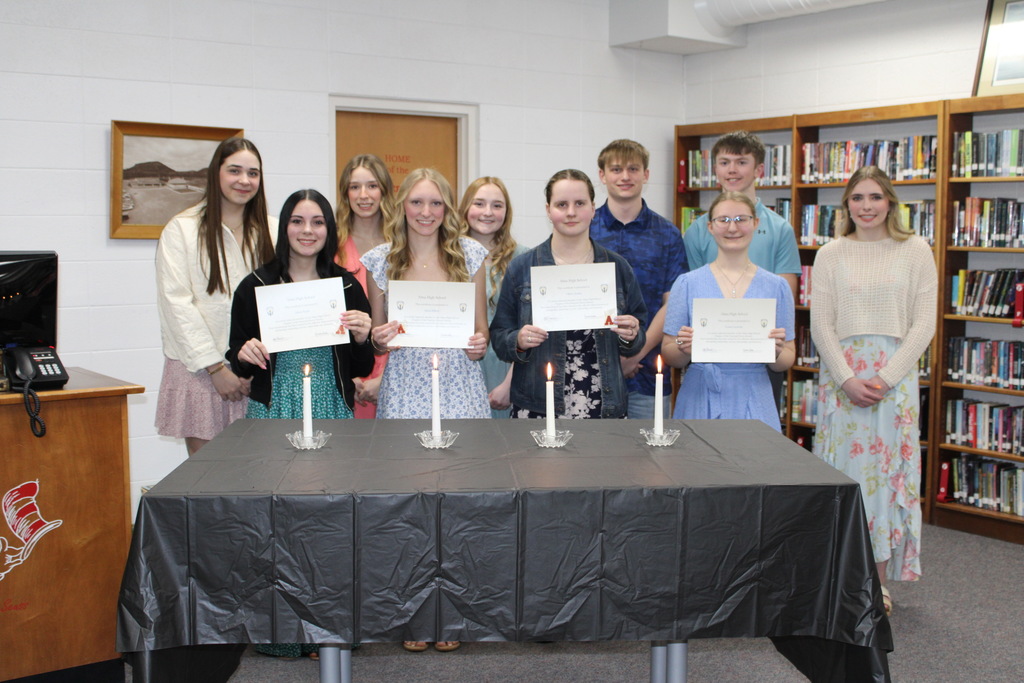 A group of students pose in a library behind a table with lit candles while several students in front hold certificates during a recognition ceremony.