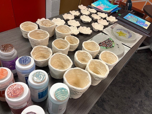 An overview of various clay projects on a table, including several tall, "coil-built" textured pots and the flower-shaped bowls. Next to them are jars of colorful ceramic glazes with fired sample lids.
