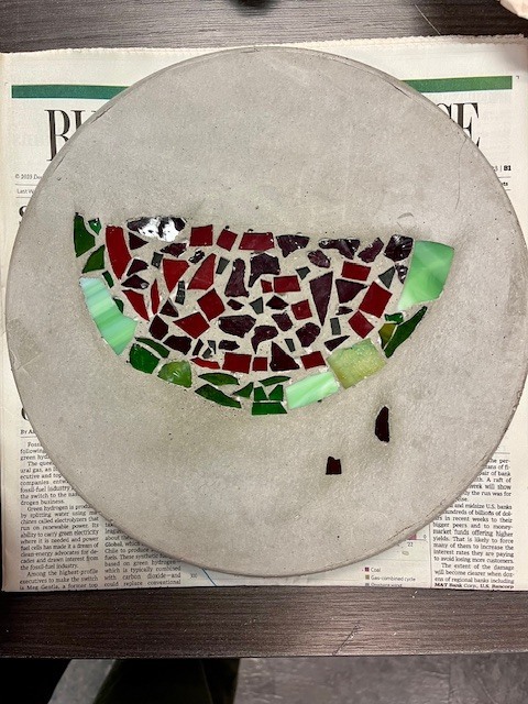 A circular concrete stepping stone with a glass mosaic of a watermelon slice. The design uses red and dark purple glass for the fruit, black for seeds, and various shades of green glass for the rind.