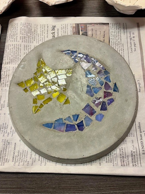 A circular concrete stepping stone featuring a glass mosaic of a crescent moon and a star. The star is made of yellow glass, and the moon is composed of iridescent blue and purple glass tiles.
