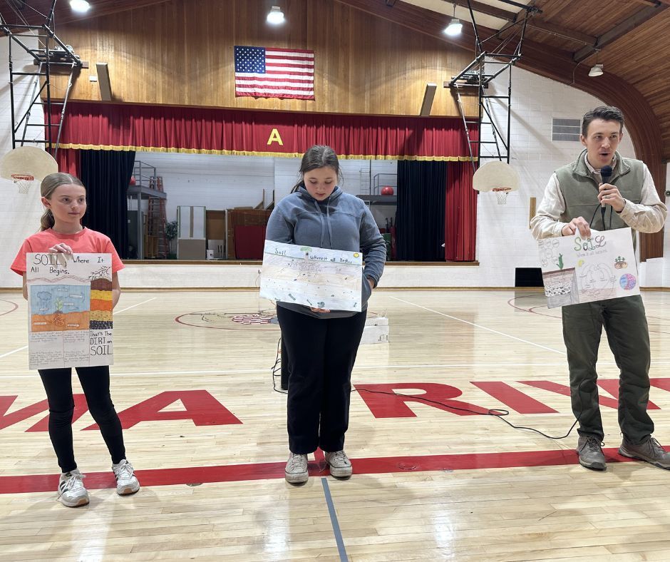 Two students and an instructor stand on a basketball court in front of a stage with a red curtain. Each person is holding a different hand-drawn educational poster about soil. An American flag hangs on the wall behind the stage.