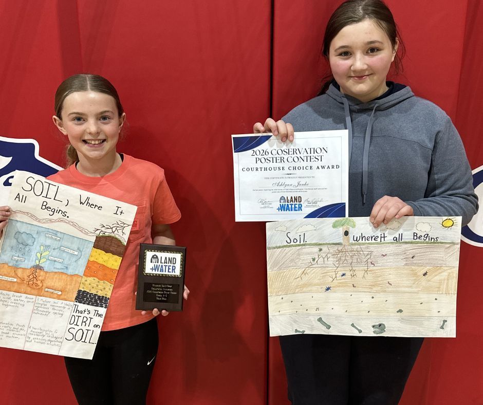 A close-up portrait of two young girls smiling in front of a red backdrop. The girl on the left holds an award plaque and a poster about soil layers. The girl on the right holds a "2026 Conservation Poster Contest" certificate and her own hand-drawn poster.