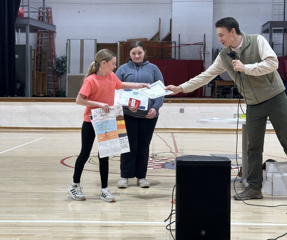 In a gymnasium, an instructor speaks into a microphone while handing a paper certificate to a student in an orange shirt. The student is holding a colorful poster about soil conservation. A second student stands nearby, watching the presentation.