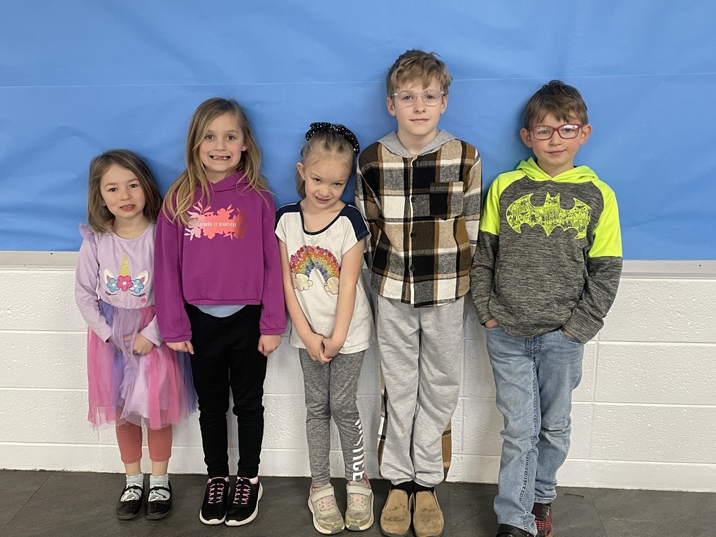 Image of elementary students smiling against a white brick wall with blue paper on the top part of it.