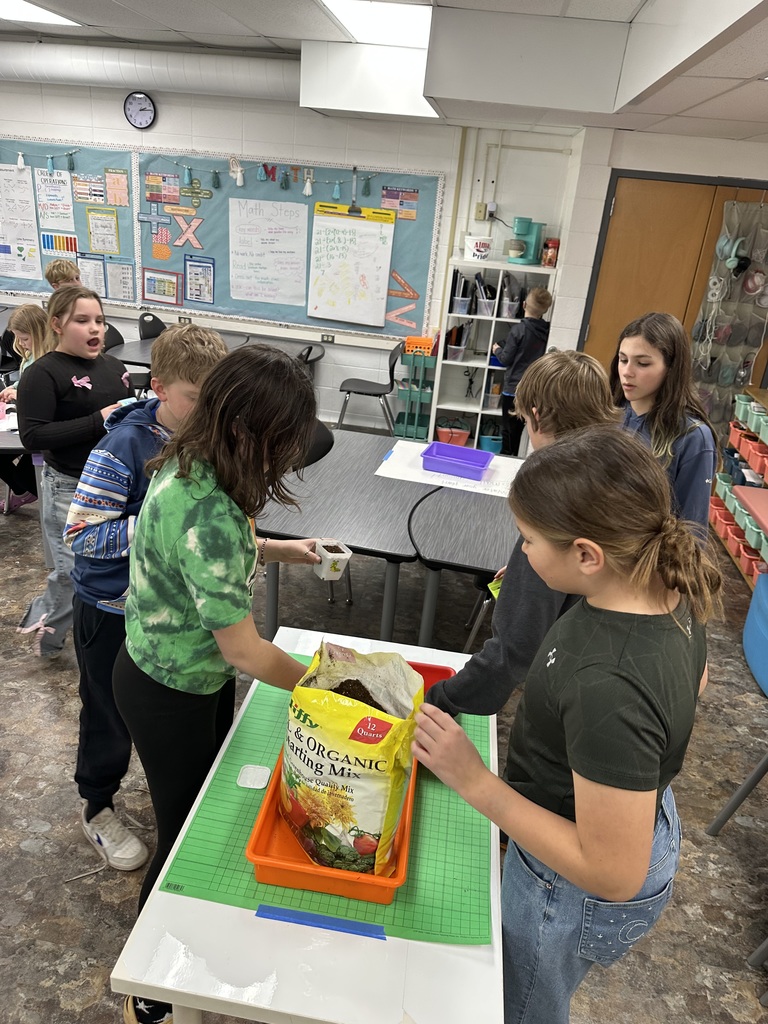 A group of elementary students gather around a table in a classroom to fill their small planters with a bag of organic starting mix soil.