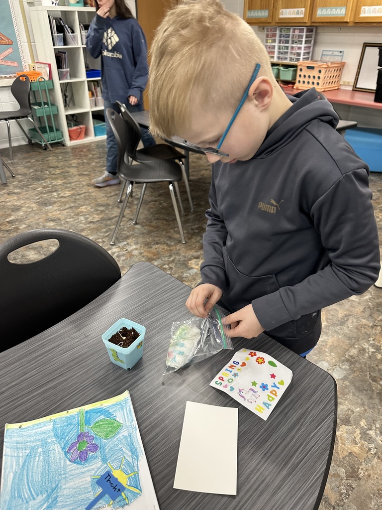 A young boy with glasses wearing a grey Puma hoodie carefully places seeds into a small blue planter at a classroom desk.