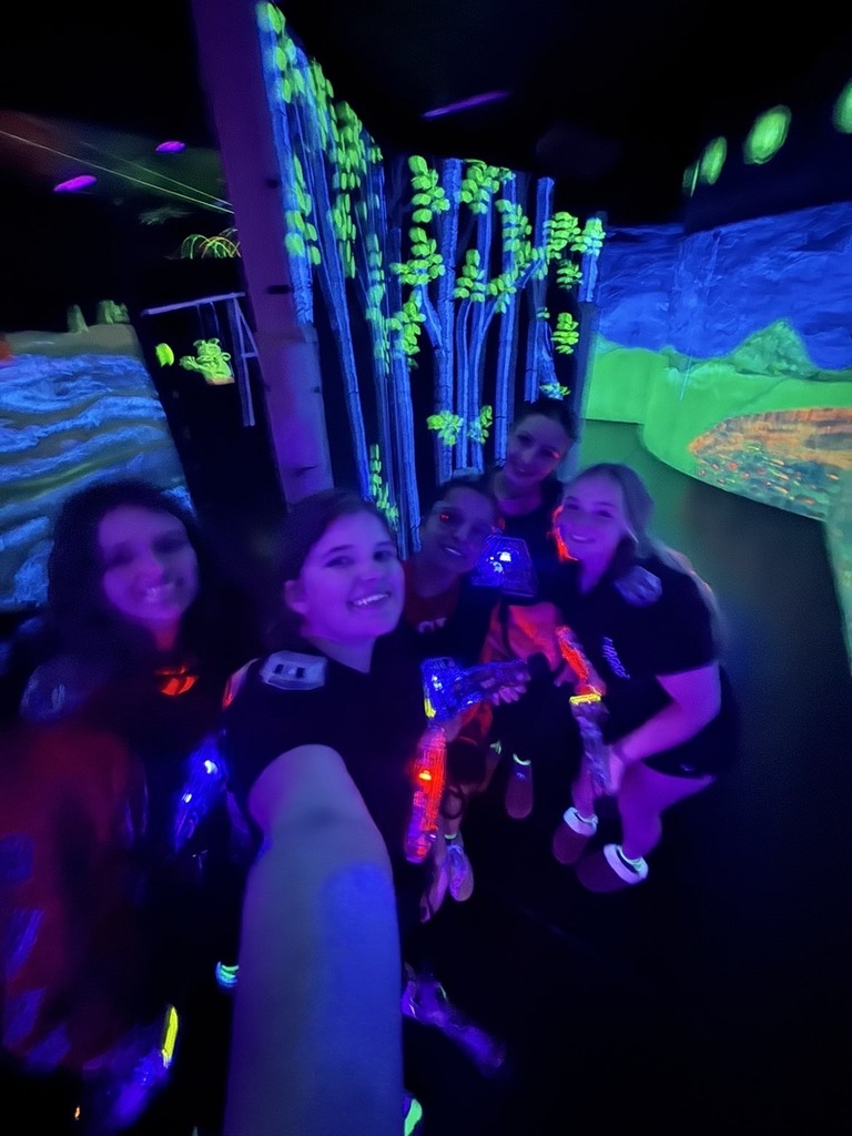 A group of five friends taking a selfie in the dark, neon-lit laser tag arena, holding glowing laser tag guns and wearing bright, colorful gear. 