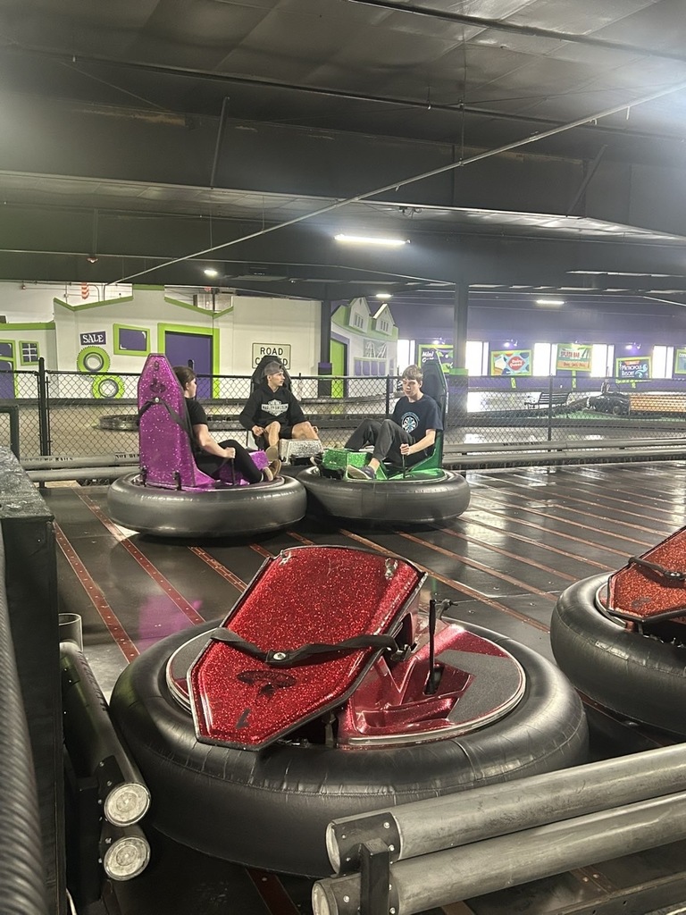 Three teens sit in colorful bumper cars on an indoor spinning bumper car track, while empty red bumper cars are parked in the foreground. The arcade-style space has black ceilings, bright lights, and green-and-purple building-themed decorations in the background. 
