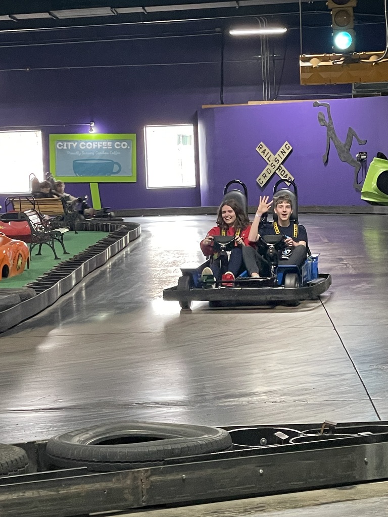 Two teenagers smiling while driving a go-kart together on an indoor track, with one waving at the camera and colorful walls and signs in the background. 