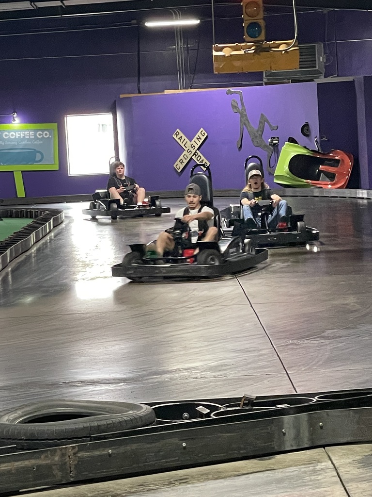 Three teenagers go-kart racing on an indoor track, focused on driving as they round a turn with purple walls and track barriers around them . 