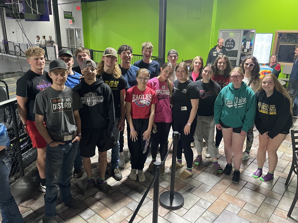 A group of about 18–20 teenagers and young adults pose together indoors, smiling at the camera. They stand in a tiled area with bright green walls, near a railing and a “City Eats” sign in the background. Many wear casual clothing such as hoodies, T-shirts, and athletic wear, with several items showing school or sports team logos like Eagles and Wisconsin. The group stands close together in a casual, friendly pose, suggesting a team or school outing.