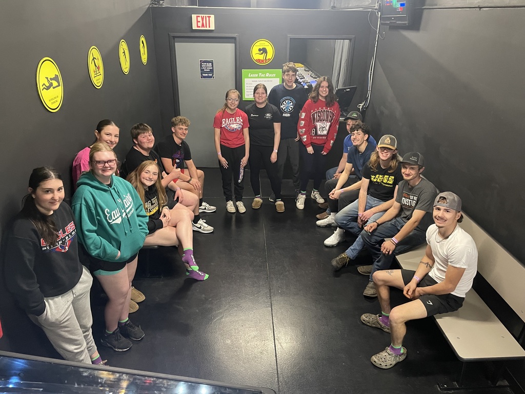 A large group of teens pose together in a dark indoor waiting area, sitting and standing along the walls before a laser tag game. Yellow  safety signs, an exit door, and a "Laser Tag Rules" sign are visible in the background. 