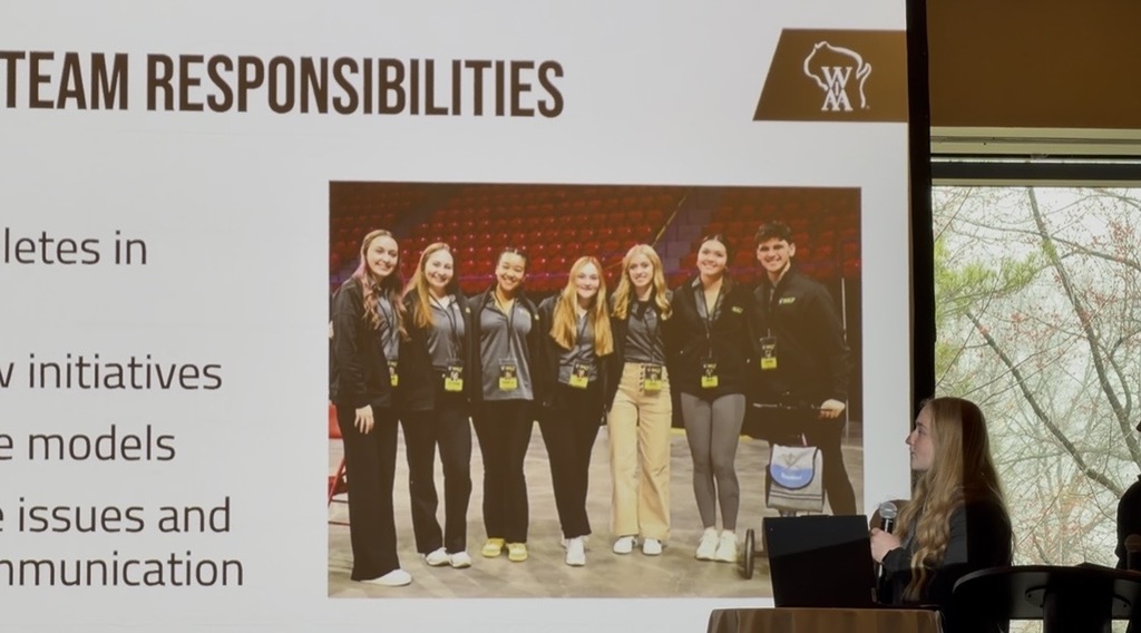 A student gives a presentation while holding a microphone and looking at a projected slide titled “Team Responsibilities.” The slide includes bullet points (partially visible) and a photo of a group of students standing together in matching outfits with event badges. A Wisconsin state athletics logo appears in the top corner of the slide. The presentation takes place in a room with large windows visible to the side.