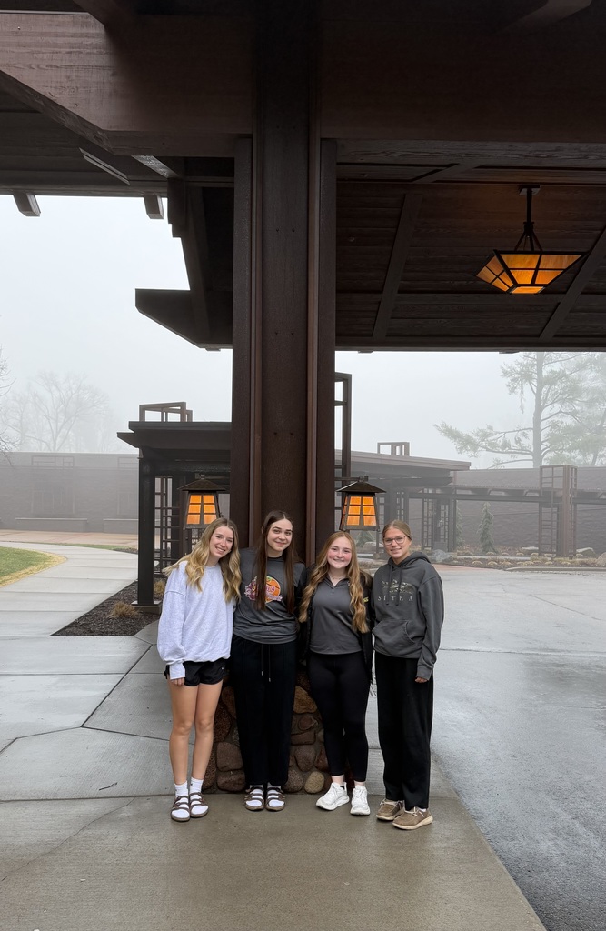 Four young women standing together under a large wooden overhang on a foggy day. They are smiling and posing in front of a lantern-style light fixture and a building with wood and stone architectural elements. The ground is wet, and trees are faintly visible in the misty background.