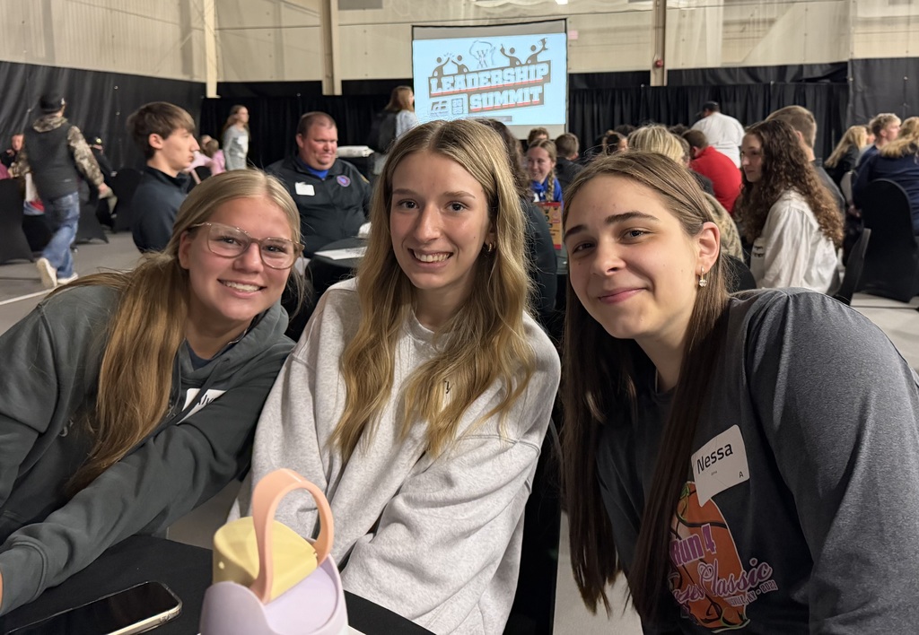 Three teenage girls smile at the camera while sitting together at a table during an indoor event. They are in a large hall with other attendees seated in the background, and a screen behind them displays “Leadership Summit.” A water bottle and a phone are on the table in front of them.