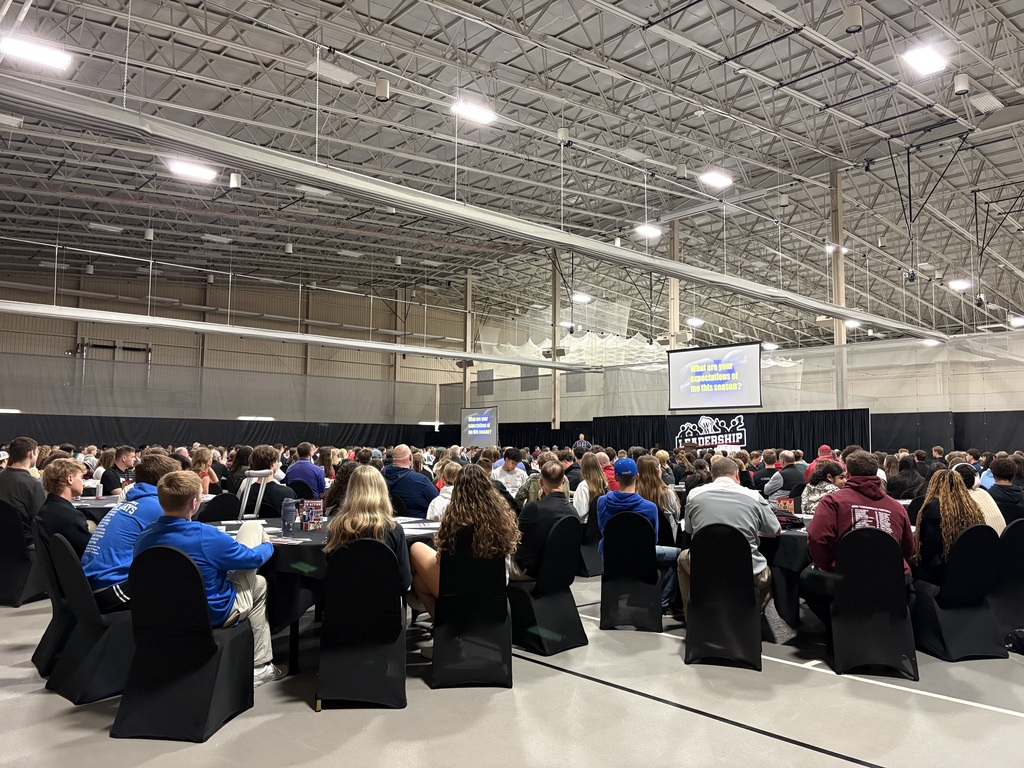 A large group of people seated at round tables in a spacious indoor venue with high industrial ceilings. At the front of the room, a stage with a large screen displays a presentation slide that reads, “What are your expectations of me this season?” The audience appears to be listening to a speaker during a leadership or team event.