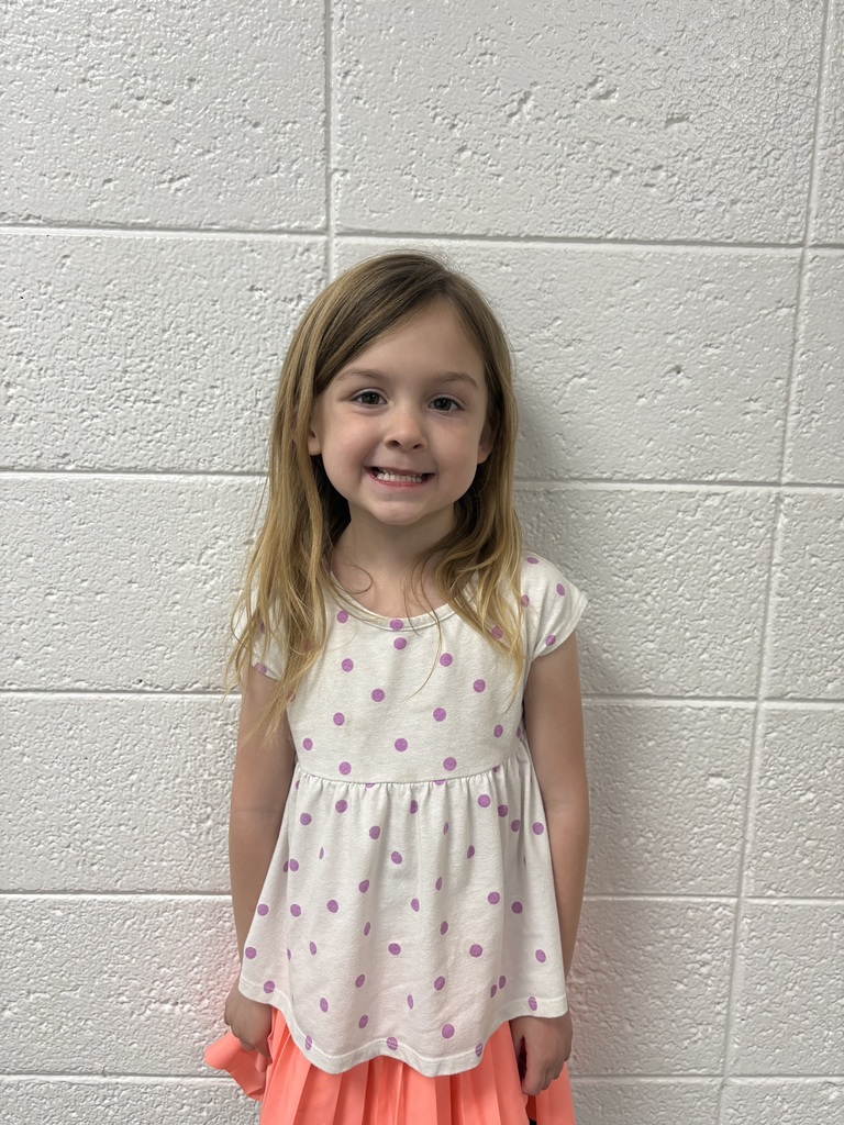 A young girl stands smiling in front of a white cinderblock wall. She has long light brown hair and is wearing a white shirt with purple polka dots and a bright coral skirt. She faces the camera with a cheerful expression, her hands relaxed at her sides.