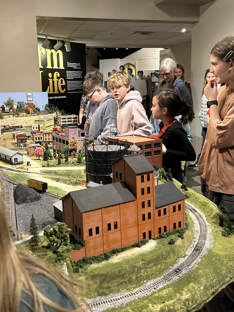 A group of fourth  grader looking at a minature train track. 