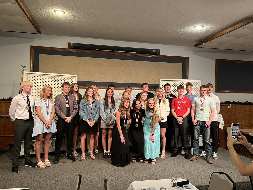 A large group of high school students stand together on a small stage at an awards event, many wearing medals around their necks. They are dressed in semi-formal attire such as dresses, button-down shirts, and slacks. The group smiles and faces the camera, with a banquet-style room setting visible, including tables and a person in the foreground holding up a phone to take a picture.