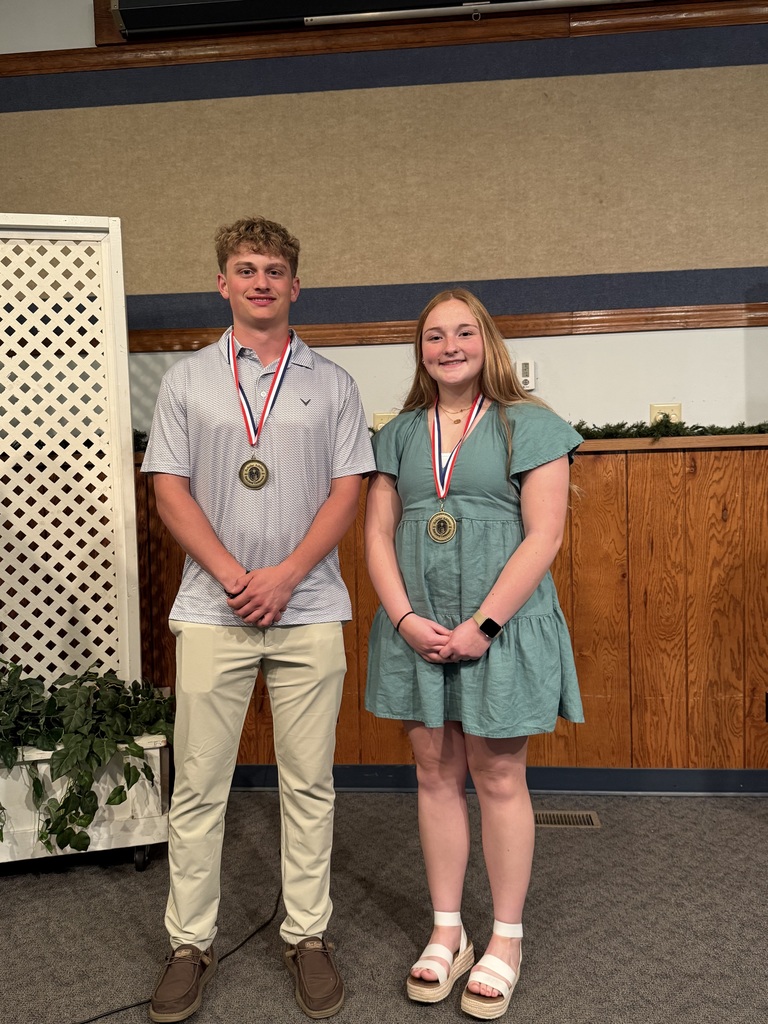Two high school students stand side by side on a stage, each wearing a medal on a ribbon around their neck. The student on the left wears a light-colored polo shirt and khaki pants, while the student on the right wears a green dress and platform sandals. Both face the camera and smile, standing in front of a wood-paneled wall with decorative lattice panels.