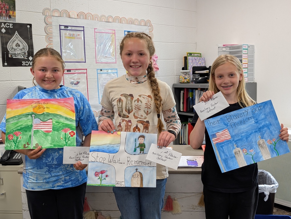 Three smiling elementary school girls stand side-by-side in a classroom, proudly holding their award-winning colorful drawings and small white envelopes from a Memorial Day poster contest. The girl on the left holds a green hill drawing and a 3rd-place note. The middle girl holds a "Stop, Wait... Remember" drawing and a 1st-place note. The girl on the right holds a blue drawing titled "Their Bravery our Freedom" and a 2nd-place note.