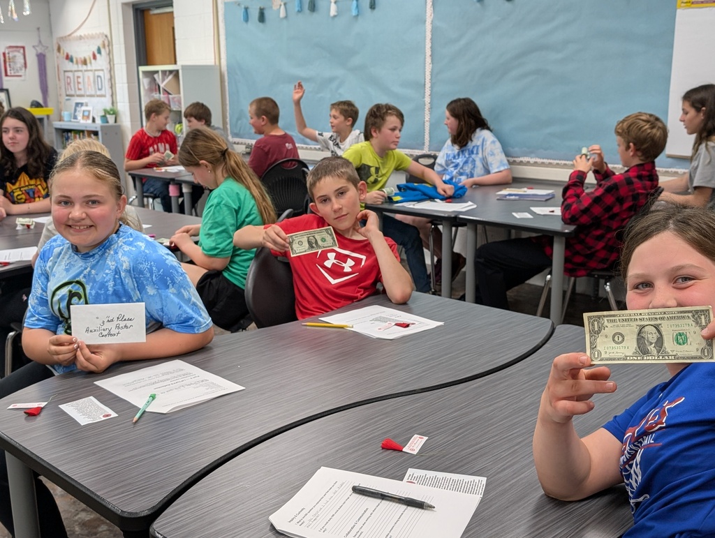 A classroom setting where elementary-aged students sit at curved gray desks. In the foreground, a girl in a blue marbled shirt smiles while holding a small white envelope that reads "3rd Place Auxiliary Poster Contest." Next to her, a boy in a red Under Armour shirt and another girl in a blue shirt hold up one-dollar bills. In the background, other children interact at their desks under a light blue bulletin board.
