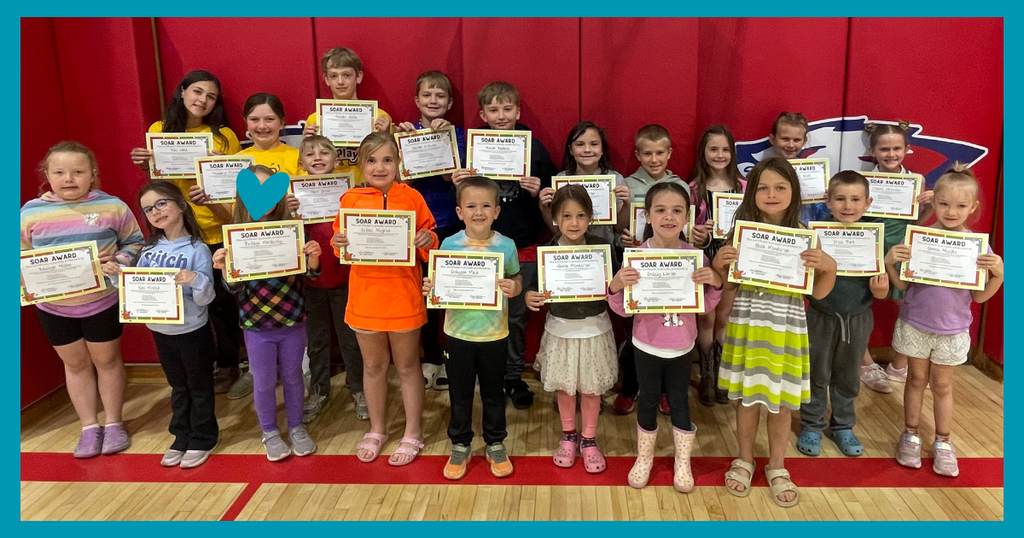 A wide group photograph of approximately twenty elementary school students standing together in a gymnasium. They are arranged in two rows against a vibrant red wall, and every student is smiling while holding up a "SOAR Award" certificate. One student's face is covered with a blue heart graphic.
