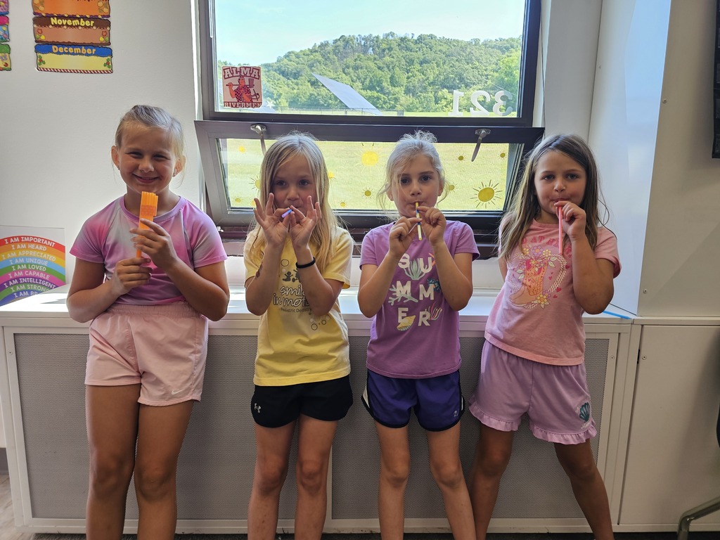 Four smiling young girls pose together in a bright room in front of a large window. They are wearing casual summer clothes—t-shirts and shorts—and holding small colorful objects like whistles or straws. A scenic green hill is visible through the window behind them.